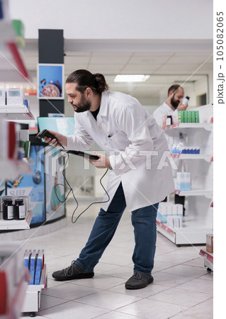 Drugstore worker looking at drugs packages while scanning supplements products using store scanner typing bar code on tablet. Pharmacist checks the expiration date on the medication boxes in pharmacy 105082065