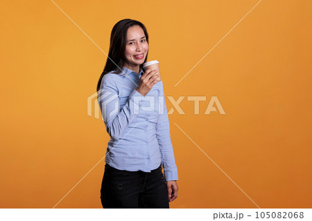 Asian woman holding cup of coffee drinking hot beverage enjoying work break before start to shoot in studio. Excited fashionable model with brunette hair standing over yellow background Asian woman holding cup of coffee drinking hot beverage enjoying work break before start to shoot in studio. Excited fashionable model with brunette hair standing over yellow background 105082068