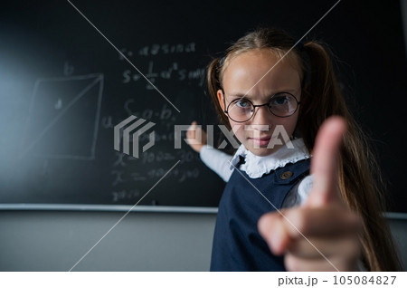 Portrait of a caucasian girl in glasses in the classroom. The schoolgirl writes the formula with chalk on the blackboard and looks into the camera. Portrait of a caucasian girl in glasses in the classroom. The schoolgirl writes the formula with chalk on the blackboard and looks into the camera. 105084827