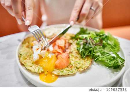Woman cuts Zucchini pancakes, with avocado, cheese cream, salmon and egg . Healthy breakfast, protein. Restaurant dish. Breakfast in cafe on a sunny morning, on the summer veranda 105087558