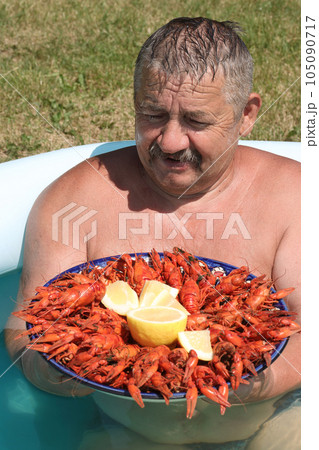 Joyful mustachioed man sits in the pool. He holds a large dish with boiled crayfish in his hands. Crayfish are decorated with lemon slices. Close-up. Joyful mustachioed man sits in the pool. He holds a large dish with boiled crayfish in his hands. Crayfish are decorated with lemon slices. Close-up. 105090717