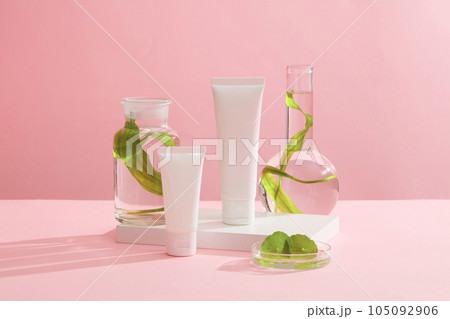 Mockup scene for advertising cosmetic with natural extract. Two plastic bottle unbranded decorated with lab glassware filled with seaweed and centella leaves on a pink background. 105092906