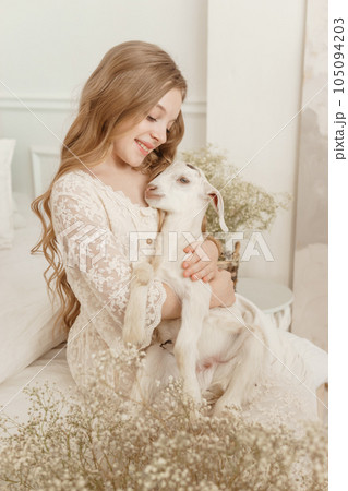 A blonde girl with long hair is sitting on the couch with a goat at home. The concept of animal care. 105094203