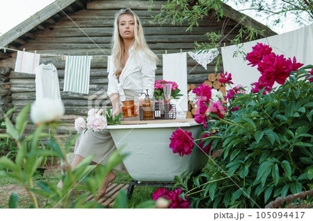 A woman is sitting on a cast-iron bathtub in the courtyard of a country house next to a bush of flowering peonies. The concept of summer, country life, a bathroom on the street in a blooming garden in 105094417