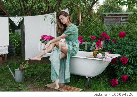 A woman is sitting on a cast-iron bathtub in the courtyard of a country house next to a bush of flowering peonies. The concept of summer, country life, a bathroom on the street in a blooming garden in A woman is sitting on a cast-iron bathtub in the courtyard of a country house next to a bush of flowering peonies. The concept of summer, country life, a bathroom on the street in a blooming garden in 105094526