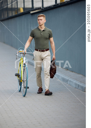 Full-length image of stylish, young businessman, employee in smart casual clothes walking along the street with bicycle. On way to office 105095603