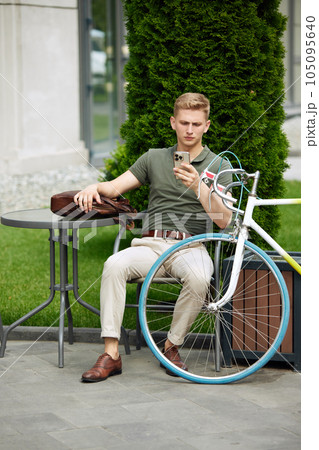 Stylish, handsome, serious businessman sitting on bench and reading news, mails on mobile phone. Going to work on bike 105095640