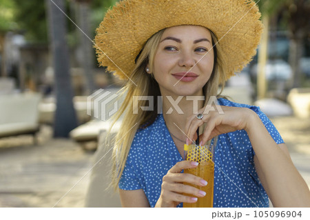 Fashion young woman drinking cocktail in beach bar. Cheerful attractive blonde woman in a dress smiles and drinks a cocktail outdoors. Happy woman enjoying summer time at the beach 105096904