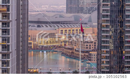 Aerial view of Dubai Fountain in downtown with palms in park next to shopping mall and souq day to night timelapse, UAE 105102563