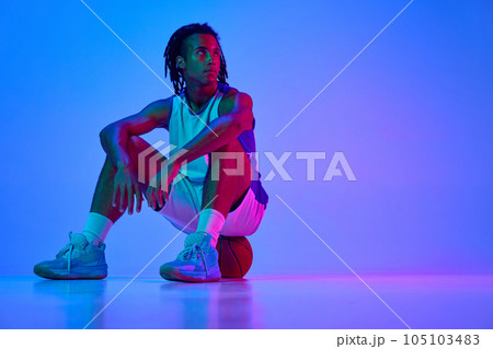 Portrait of young guy, basketball player in uniform, sitting on floor, posing against gradient blue background in neon lights 105103483