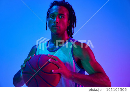 Young man with dreads, basketball player in uniforma posing with ball against gradient blue background in neon lights 105103506