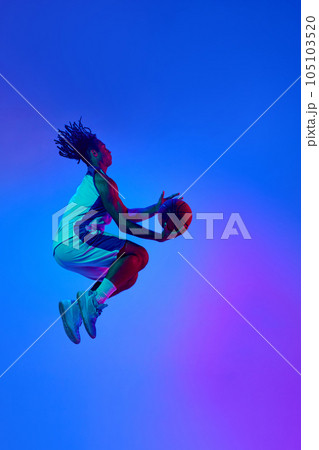 Full-length image of young professional basketball player in motion, training, playing against gradient blue background in neon lights 105103520