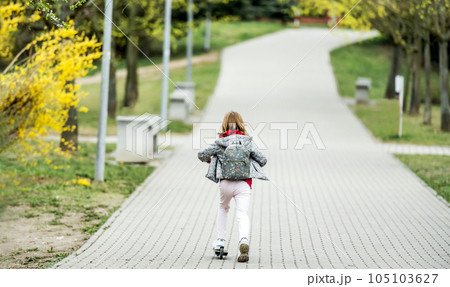 Little cute girl riding a scooter on a path in the park, back view Little cute girl riding a scooter on a path in the park, back view 105103627
