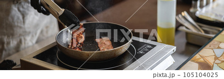 Close-up view of chef in uniform stir frying bacon in pan for cooking dish in restaurant kitchen 105104025
