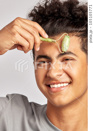 Young handsome smiling guy using jade de-puffing face roller to keep his skin soft and smooth. Swarthy man with black curly hair maintains his natural beauty with a skincare routine. Men's self-care. Young handsome smiling guy using jade de-puffing face roller to keep his skin soft and smooth. Swarthy man with black curly hair maintains his natural beauty with a skincare routine. Men's self-care. 105105771