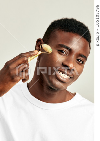Young handsome black guy using jade de-puffing face roller to keep his skin soft and smooth. African American millennial man maintains his natural beauty with a skincare routine. Men's self-care. Young handsome black guy using jade de-puffing face roller to keep his skin soft and smooth. African American millennial man maintains his natural beauty with a skincare routine. Men's self-care. 105105796