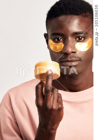 Young handsome black guy applying cosmetic hydrogel patches under his eyes from a case in his hand. African American millennial man practices skin care routine to keep healthy and youthful looking. 105106030