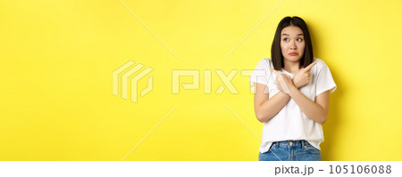 Indecisive asian girl need help with choice, pointing fingers sideways and looking confused, standing over yellow background 105106088