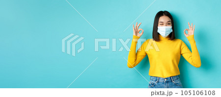 Covid-19, social distancing and pandemic concept. Cheerful asian girl in medical mask showing okay gestures, winking at camera, showing approval, standing over blue background 105106108