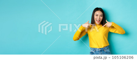 Beauty and fashion concept. Confident and sassy asian woman pointing at herself, smiling cheeky at camera, being professional, standing over blue background 105106206