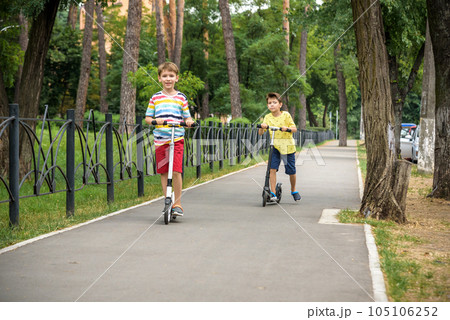 Two attractive European boys brothers, wearing red and white checkered shirts, standing on scooters in the park. They laughing, smiling, hugging and having fun. Active leisure time with kids 105106252