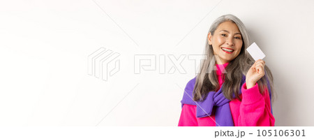 Shopping concept. Stylish asian senior woman smiling and showing plastic credit card, paying contactless, standing over white background Shopping concept. Stylish asian senior woman smiling and showing plastic credit card, paying contactless, standing over white background 105106301