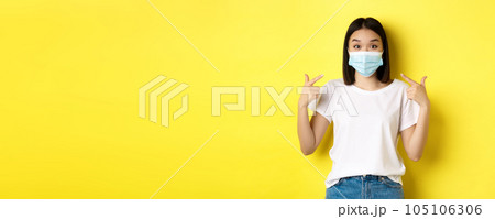 Covid-19, quarantine and social distancing concept. Young asian woman in white t-shirt, pointing at her medical mask from coronavirus, yellow background Covid-19, quarantine and social distancing concept. Young asian woman in white t-shirt, pointing at her medical mask from coronavirus, yellow background 105106306