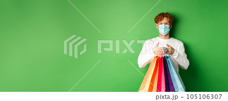 Pandemic and lifestyle concept. Cheerful redhead man going shopping in store, wearing medical mask and holding bags, standing over green background Pandemic and lifestyle concept. Cheerful redhead man going shopping in store, wearing medical mask and holding bags, standing over green background 105106307