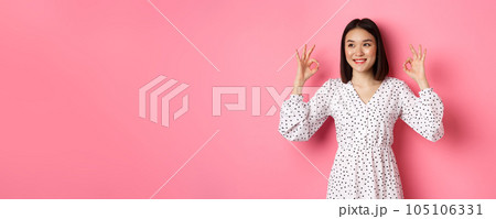 Pretty young asian woman in dress showing okay sign, praising and showing approval, looking satisfied, standing against pink background Pretty young asian woman in dress showing okay sign, praising and showing approval, looking satisfied, standing against pink background 105106331