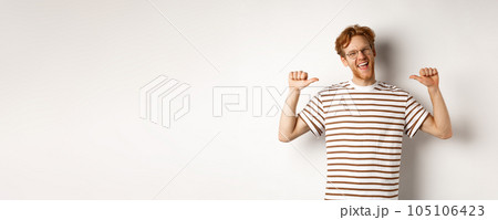 Cheerful and funny redhead man with beard, wearing glasses, laughing and pointing at himself, standing over white background 105106423