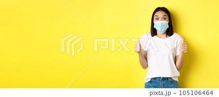 Covid-19, quarantine and social distancing concept. Cheerful asian woman in medical mask and white t-shirt showing thumbs up in approval, praise good deal, yellow background 105106464