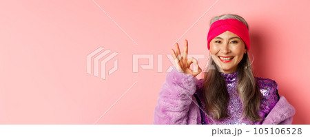 Fashion. Close-up of asian old lady in stylish disco outfit showing OK sign, approve and like something, smiling pleased, pink background 105106528