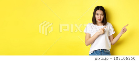 Beauty and fashion concept. Beautiful asian woman in white t-shirt pointing fingers right, demonstrate logo standing over yellow background 105106550