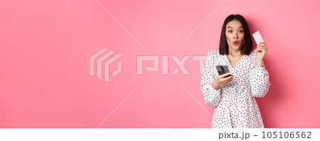 Attractive young asian woman order online, holding credit card and mobile phone, making internet purchase, standing happy over pink background 105106562