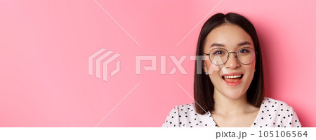 Beauty and lifestyle concept. Close-up of cute asian female model wearing trendy glasses, smiling happy at camera, standing on pink background 105106564
