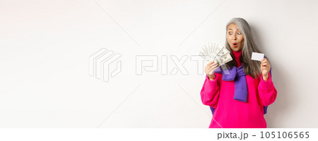 Shopping concept. Lucky asian senior woman looking amazed at money and showing plastic credit card, standing over white background 105106565