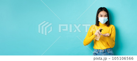 Covid-19, social distancing and pandemic concept. Smiling asian woman in medical mask using preventive measures from coronavirus, using hand sanitizer, standing over blue background 105106566