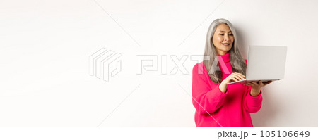 Senior asian woman working freelance, using laptop and smiling, standing over white background 105106649