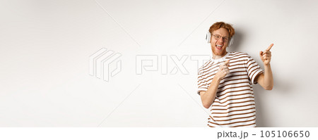 Technology concept. Happy redhead man listening music in headphones and dancing and pointing fingers right, standing over white background 105106650