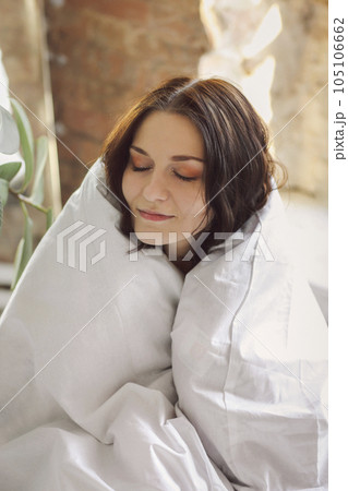 Portrait of young beautiful happy woman, sitting wrapped in duvet with eyes closed 105106662