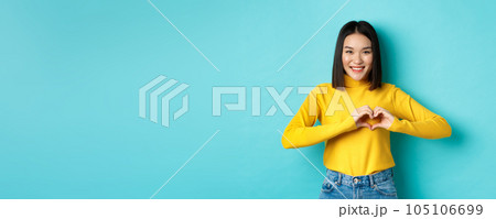 Valentines day and romance concept. Beautiful asian woman show I love you, heart gesture and smiling, standing against blue background 105106699