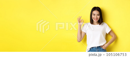 Confident asian woman smiling and showing OK sign, approve and praise good offer, standing in white t-shirt over yellow background Confident asian woman smiling and showing OK sign, approve and praise good offer, standing in white t-shirt over yellow background 105106753