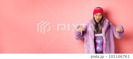 Fashion and shopping concept. Funky asian senior woman in trendy faux fur coat and disco dress, looking and pointing down, standing over pink background Fashion and shopping concept. Funky asian senior woman in trendy faux fur coat and disco dress, looking and pointing down, standing over pink background 105106761