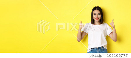 Pretty young asian woman in white t-shirt, showing thumbs up and smiling, praise good offer, recommend product, standing satisfied over yellow background Pretty young asian woman in white t-shirt, showing thumbs up and smiling, praise good offer, recommend product, standing satisfied over yellow background 105106777