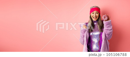Cheerful asian senior woman with grey hair, wearing party glitter dress, showing delicious donut and smiling happy, eating sweet food, standing over pink background Cheerful asian senior woman with grey hair, wearing party glitter dress, showing delicious donut and smiling happy, eating sweet food, standing over pink background 105106788