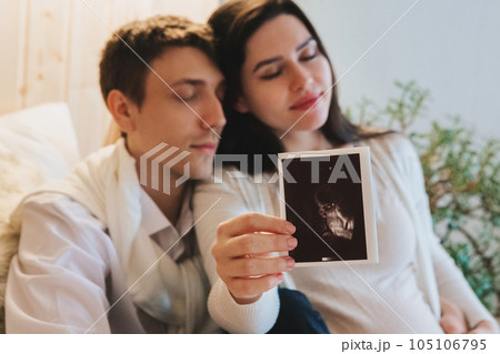 Young happy pregnant woman holding pregnancy ultrasound, showing sonogram picture to pleasantly surprised husband crying from happiness. Family couple waiting for baby Young happy pregnant woman holding pregnancy ultrasound, showing sonogram picture to pleasantly surprised husband crying from happiness. Family couple waiting for baby 105106795