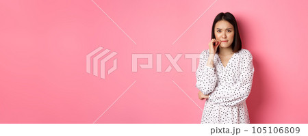 Brunette asian woman in dress looking displeased, frowning and zipping mouth, seal lips with promise, standing over pink background 105106809