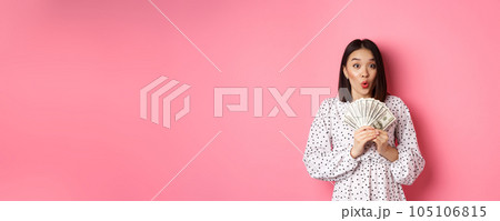 Shopping concept. Excited asian woman holding money, gasping amazed and staring at camera, standing over pink background 105106815