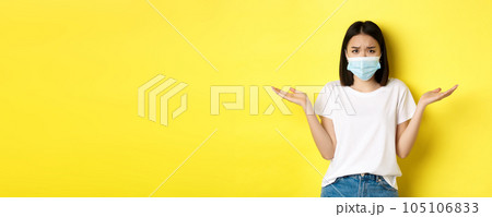 Covid-19, quarantine and social distancing concept. Confused asian woman in medical mask shrugging shoulders, spread hands sideways clueless, know nothing, yellow background Covid-19, quarantine and social distancing concept. Confused asian woman in medical mask shrugging shoulders, spread hands sideways clueless, know nothing, yellow background 105106833