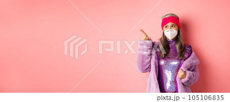Covid and fashion concept. Stylish asian senior woman checking out special deal, wearing disco dress and face mask, pointing finger left at copy space, pink background 105106835